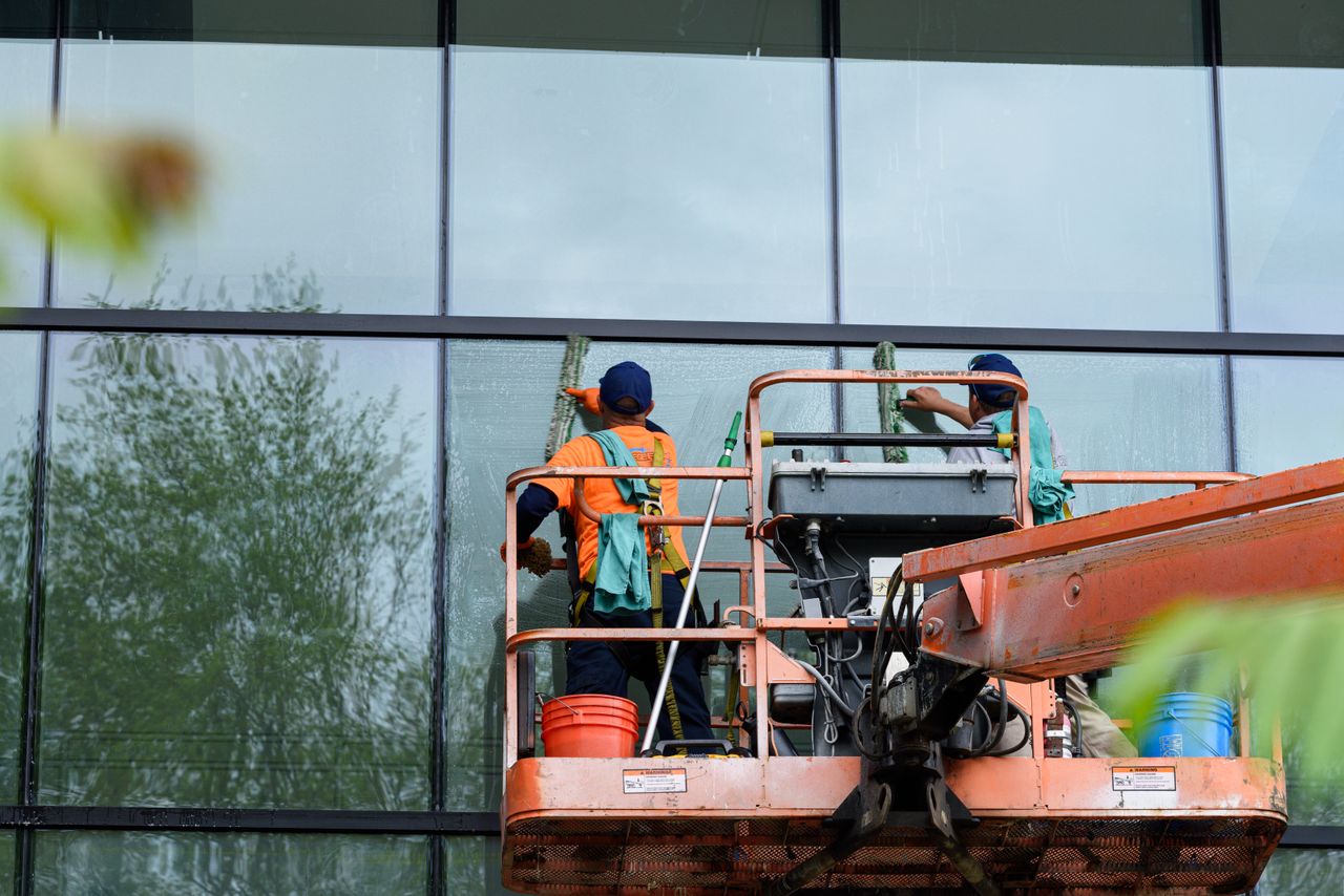 about-02 Two workers cleaning windows on a glass building facade using a lift platform, enhancing building aesthetics.